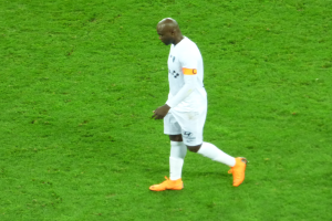 Ein Fussballspieler in einem wei├čen T-Shirt, wei├čen Shorts und orangenen Schuhen geht ├╝ber ein saftig gr├╝nes Feld.