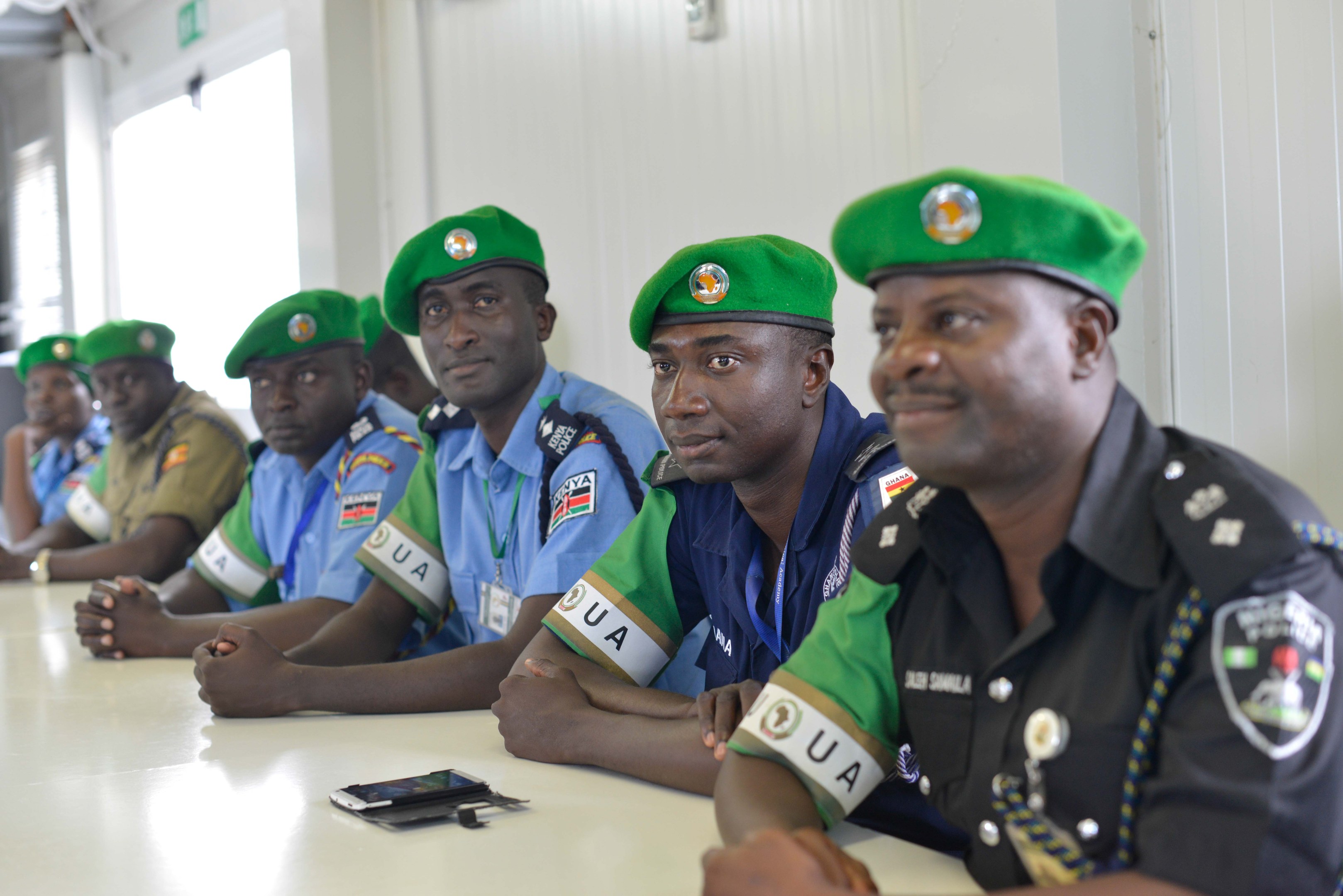 Gruppe von Polizeibeamten bei einer Schulung an einem Tisch sitzend, mit Mobiltelefonen und anderen Gegenständen auf dem Tisch, in einem Raum mit Fenstern und Deckenbeleuchtung.
