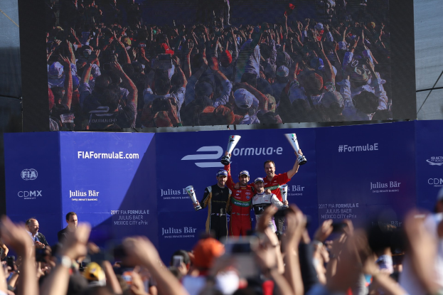 Gruppe von Menschen auf einem Formel-E-Podium in Mexiko-Stadt mit Pokalen, Sponsor-Plakaten und einem Bildschirm im Hintergrund.