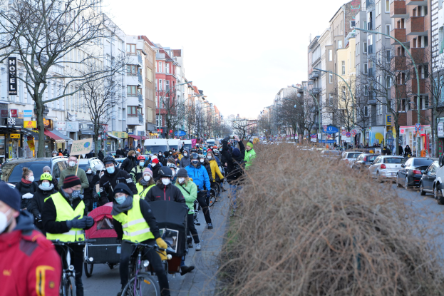 Eine große Gruppe von Menschen in Masken und Sicherheitswesten auf Fahrrädern eine straße entlang, gesäumt von Bäumen, Gebäuden mit Fenstern, Laternenmasten und Schildern, neben Fahrzeugen, trockenem Gras und einem klaren blauen Himmel.