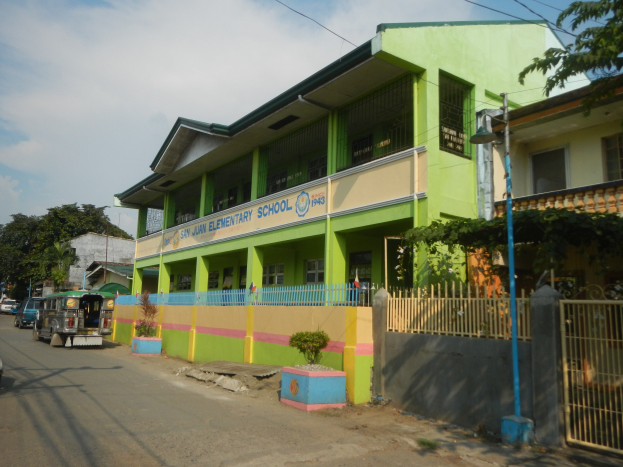 Schulgebäude mit grüner und gelber Fassade, Fahrzeuge auf der Straße, Topfpflanzen, ein Zaun, ein Tor, ein Straßenpfahl, Bäume, Stromleitungen und eine bewölkte Himmel.
