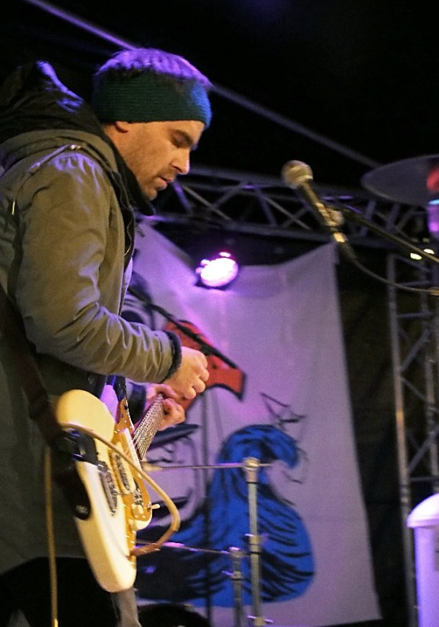 Ein Mann in einer Jacke und einer Mütze spielt eine E-Gitarre auf der Bühne, mit einem Mikrofon vor ihm, umgeben von Scheinwerfern, einem Banner und einem Schlagzeug zu seiner Rechten.