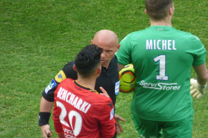 Ein Schiedsrichter, der mit zwei Fußballspielern auf einem grünen Rasen spricht, wobei einer der Spieler einen Ball hält.