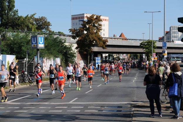 Gruppe von Menschen, die bei einem Marathon auf einer von Bäumen gesäumten Straße mit Schildern, Fahrrädern, einem Zaun, Gras, einer Brücke, Gebäuden und einem klaren blauen Himmel laufen.
