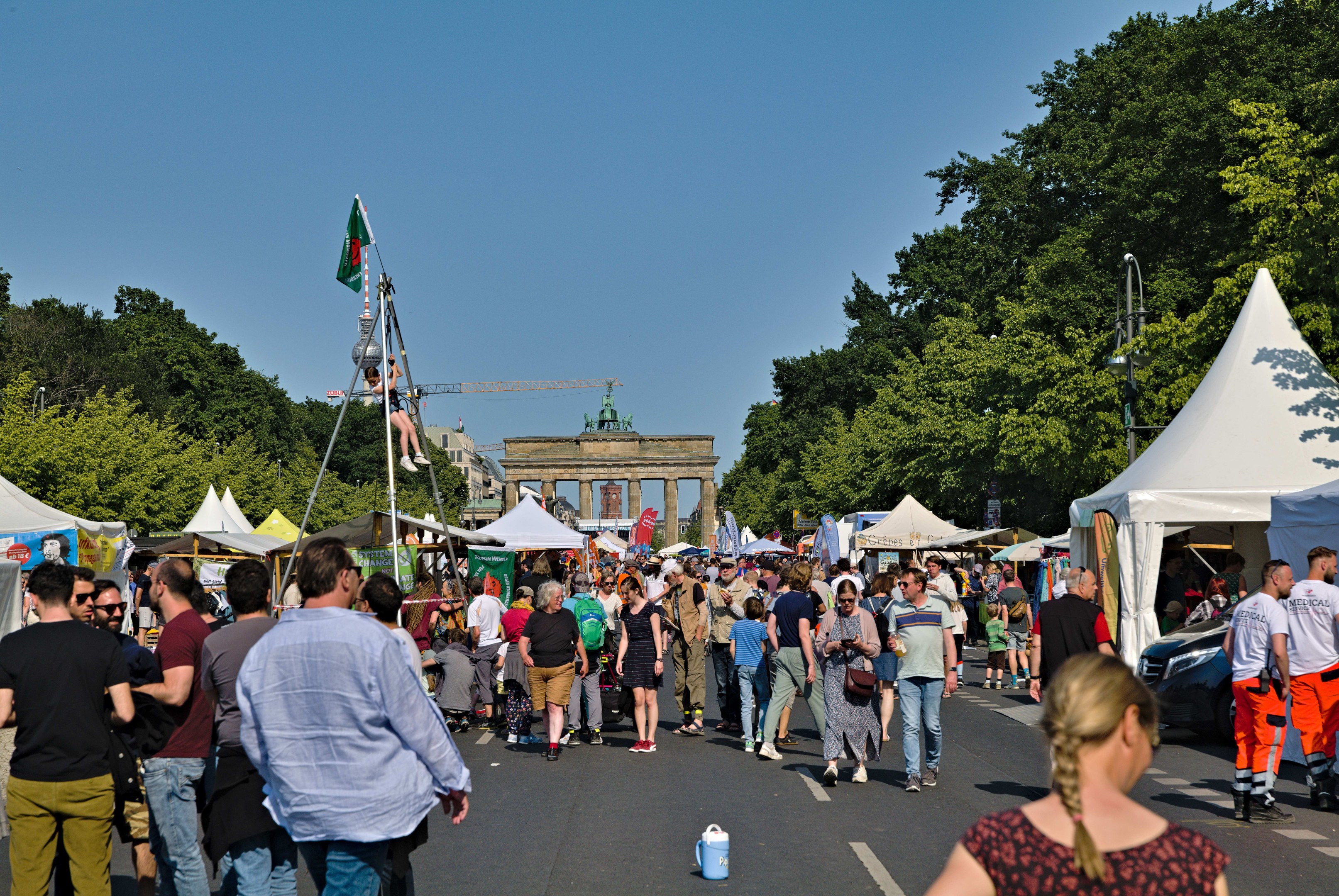 Eine Menschenmenge geht eine Straße entlang, die von Zelten, Fahrzeugen und Bäumen gesäumt ist, mit einem Bogen und einem klaren blauen Himmel im Hintergrund und Fahnen auf Stangen, die das Oktoberfest in München, Deutschland, anzeigen.