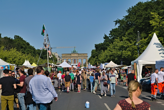 Eine Menschenmenge geht eine Straße entlang, die von Zelten, Fahrzeugen und Bäumen gesäumt ist, mit einem Bogen und einem klaren blauen Himmel im Hintergrund und Fahnen auf Stangen, die das Oktoberfest in München, Deutschland, anzeigen.