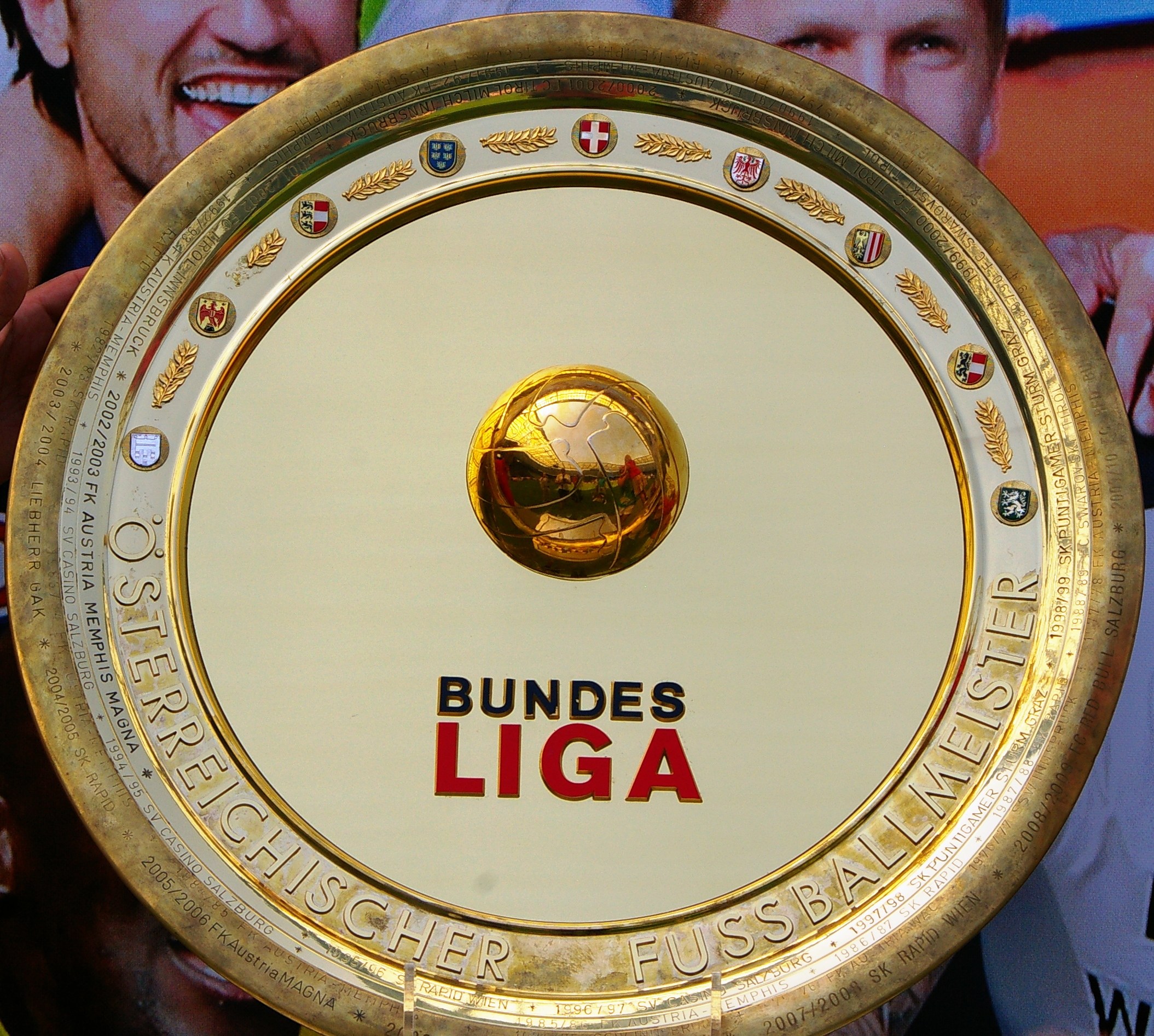 Ein Mann hält einen goldenen Schild mit der Aufschrift "Bundesliga", im Hintergrund eine Fahne mit drei Personen.