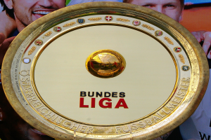 Ein Mann hält einen goldenen Schild mit der Aufschrift "Bundesliga", im Hintergrund eine Fahne mit drei Personen.