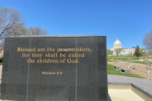Ein Gedenkstein mit der Inschrift "Gesegnet sind die Friedensstifter, denn sie werden die Kinder Gottes genannt" umgeben von Bäumen, Pflanzen, Gras, Laternenmasten und einem Gebäude unter einem klaren blauen Himmel.