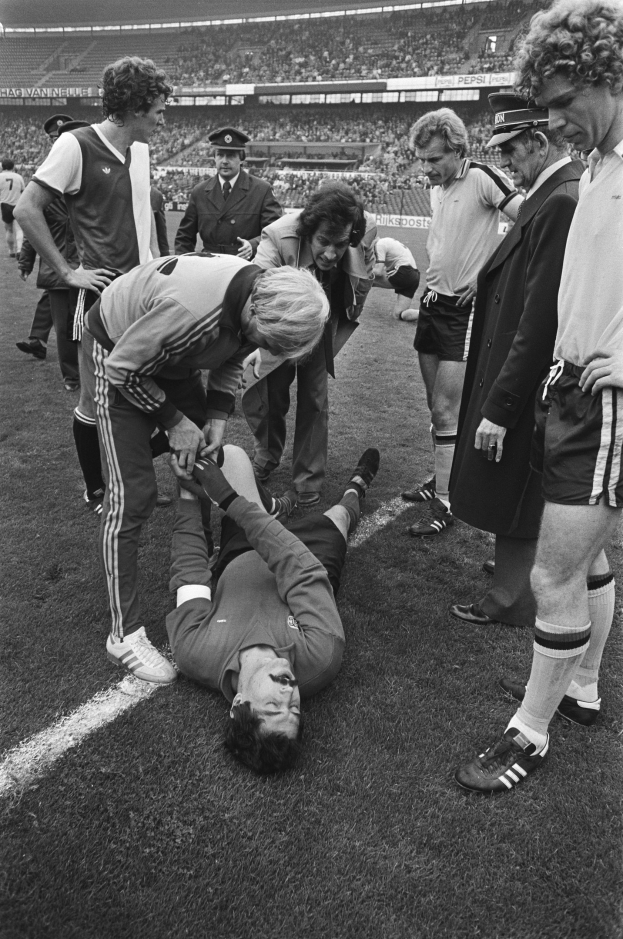 Schwarzes Bild eines Fußballspielers, der auf dem Boden liegt und von einer Gruppe Männer und einer Stadionmenge umgeben ist, der verletzt zu sein scheint.