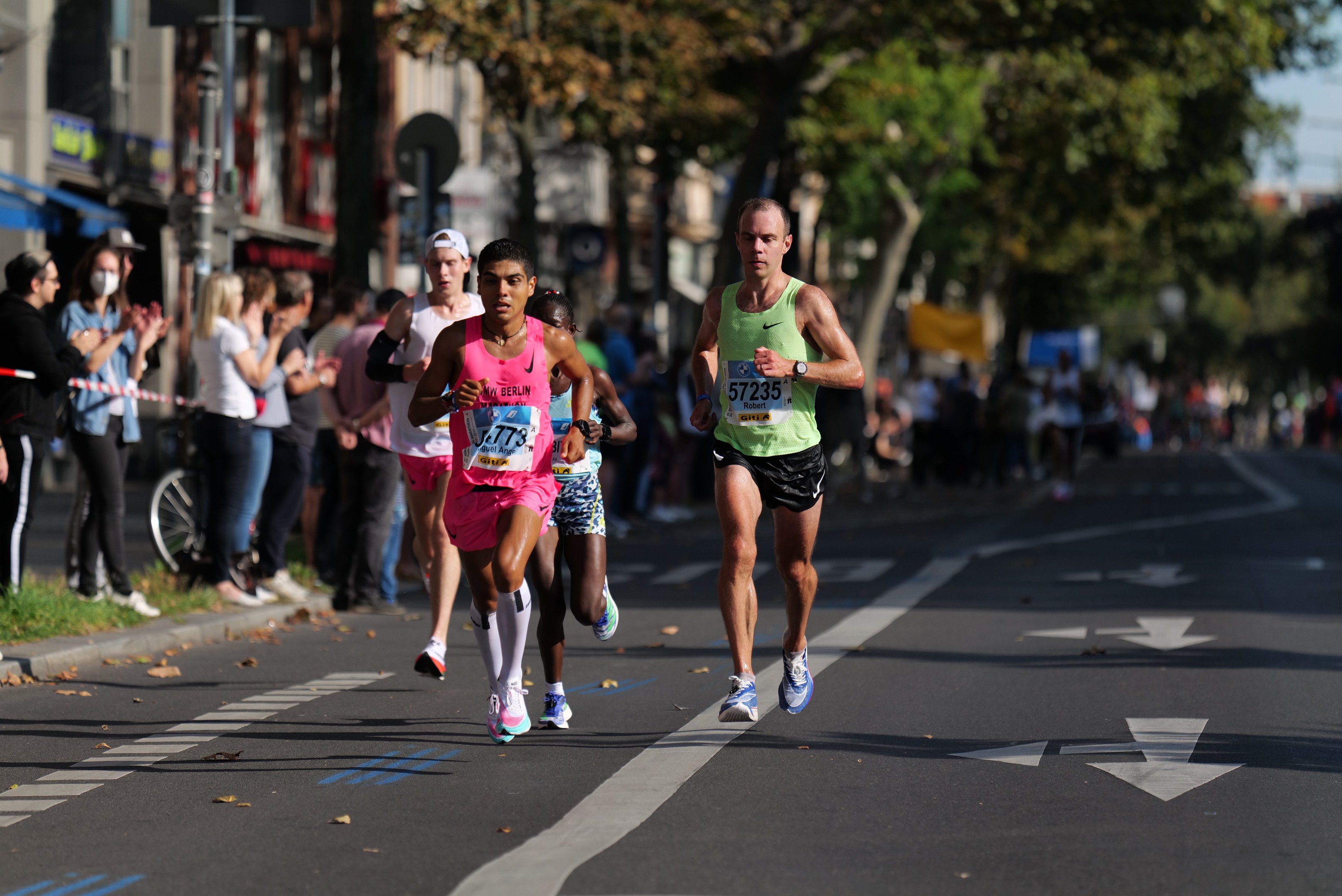 Gruppe von Menschen, die in einem Stadtmarathon laufen, mit Zuschauern auf der linken Seite, unscharfer städtischer Hintergrund mit Bäumen, Gebäuden und einem Fahrrad.