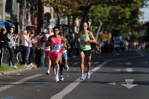 Gruppe von Menschen, die in einem Stadtmarathon laufen, mit Zuschauern auf der linken Seite, unscharfer städtischer Hintergrund mit Bäumen, Gebäuden und einem Fahrrad.