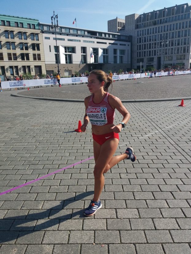 Eine Frau in einem Sportdress läuft durch eine Marathon-Strecke, die von Verkehrskegeln, einer Schleife und einem Banner markiert ist, mit Zuschauern, Straßenlaternen mit Flaggen, Gebäuden und einem bewölkten Himmel im Hintergrund.