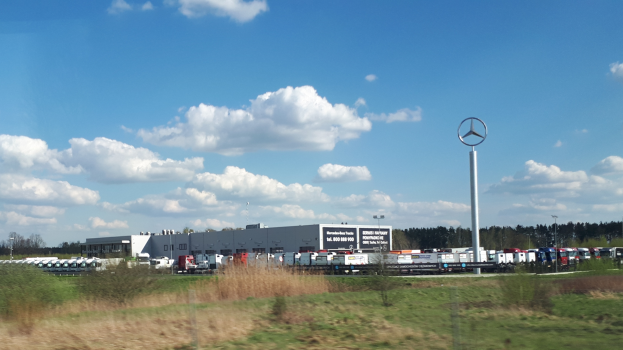 Ein Mercedes-Benz-Werk in Deutschland mit Fahrzeugen auf der Straße, umgeben von Gras, Pflanzen, Bäumen, Gebäuden und Pfählen, von denen einer ein Logo trägt, unter einem bewölkten Himmel.