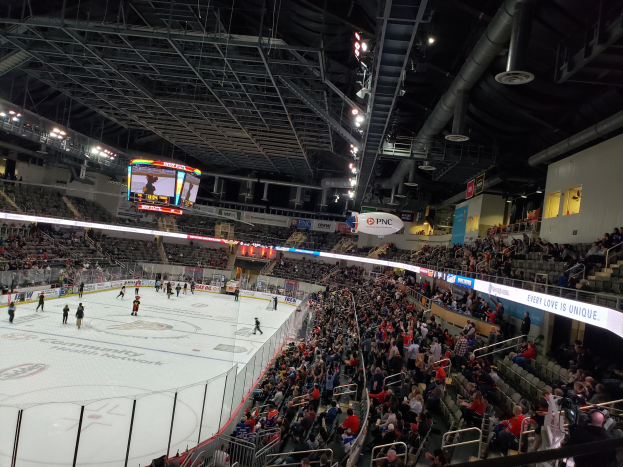 Eishockeyspiel in einer großen Arena mit sitzenden und stehenden Zuschauern in der Nähe der Eisfläche, umgeben von Bannern, Fenstern und Deckenbeleuchtung im PNC Arena.