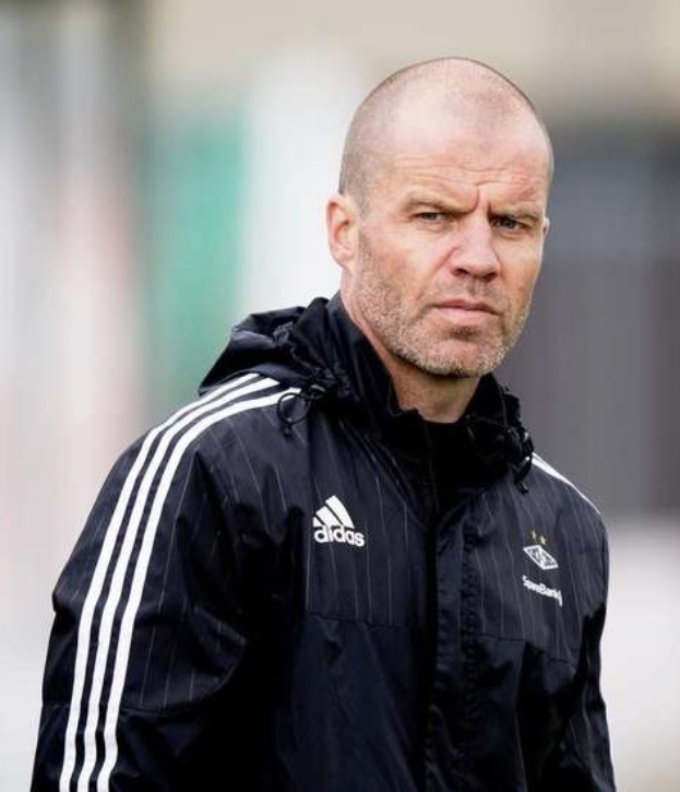 Ein kahlköpfiger Trainer in einer schwarzen Jacke steht auf einem Fußballfeld und trägt eine marineblaue Jacke mit weißen Streifen und hat die Hände verschränkt.