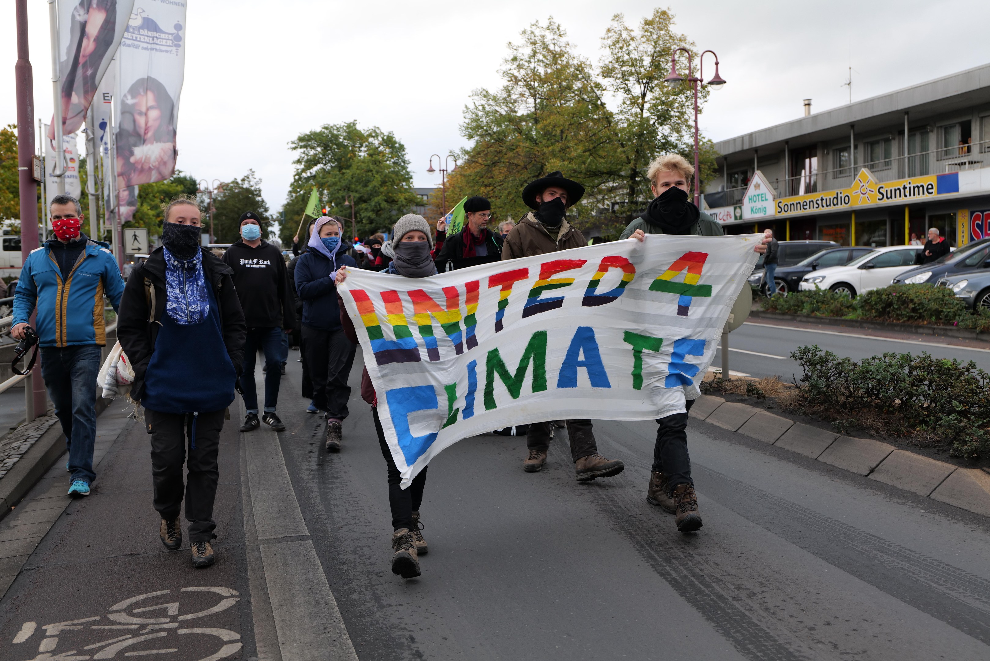 Eine Gruppe von Menschen in Masken, die eine "United 4 Climate"-Tafel tragen, geht eine Straße entlang, mit einem Gehweg, Geländer, zusätzlichen Tafeln, Bäumen, Laternen, Fahrzeugen, Gebäuden, Namensschildern und einem klaren blauen Himmel im Hintergrund.