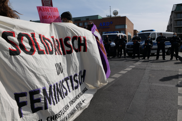 Gruppe von Menschen, die eine "Solidarität und Feminismus"-Fahne schwenken, marschiert eine Straße entlang, mit geparkten Fahrzeugen, Gebäuden, einer Schüsselantenne und einem klaren blauen Himmel im Hintergrund.