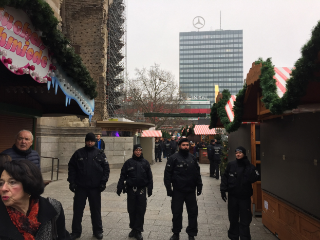 Polizisten in schwarzen Uniformen stehen vor einem geschm√∞ckten Weihnachtsmarkt in Berlin, mit festlichen Dekorationen, Gebäuden, Bäumen und einem klaren blauen Himmel im Hintergrund.