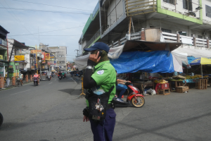 Ein Polizist in grüner und blauer Uniform und blauer Mütze steht an der Straße und spricht in ein Handy, während hinter ihm Fahrzeuge fahren und rechts von ihm Stände mit verschiedenen Gegenständen zu sehen sind, und im Hintergrund ein Gebäude mit Fenstern unter einem bewölkten Himmel.