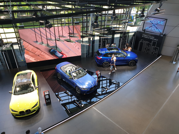 Eine Gruppe von Menschen, die auf dem Boden des BMW Museums in München stehen, mit Autos zur Linken und einem Schild mit der Aufschrift "BMW Museum München" zur Rechten, überblicken sie B├Ąume, Geb├Ąude und den Himmel durch Glasfenster.