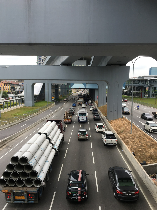 Autobahn mit starkem Verkehr unter einer Brücke, umgeben von Gras, Bäumen, Gebäuden, Polen und Straßenlaternen unter einem Himmel.