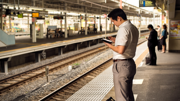 Ein Mann steht auf einem Bahnsteig, schaut auf sein Handy, umgeben von anderen Menschen mit Bahnschienen im Hintergrund und einer Tafel mit Text auf der rechten Seite.