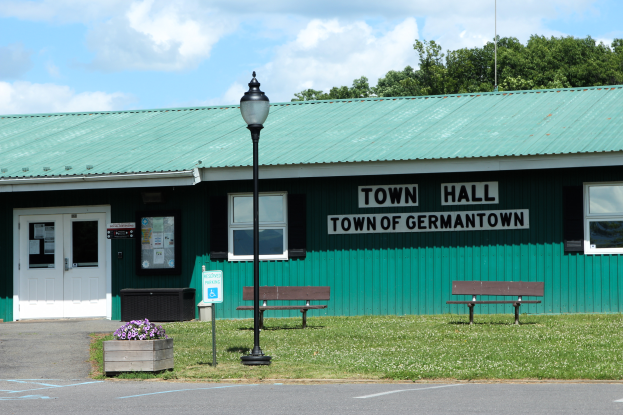 Ein Rathaus-Gebäude in Germantown, Pennsylvania, mit Fenstern, Türen und einem Dach, umgeben von Bänken, Gras, einem Straßenmast, einem Schild, einem Müllbehälter, Blumentöpfen mit Blumen, einer Baumgruppe und einem bewölkten Himmel.
