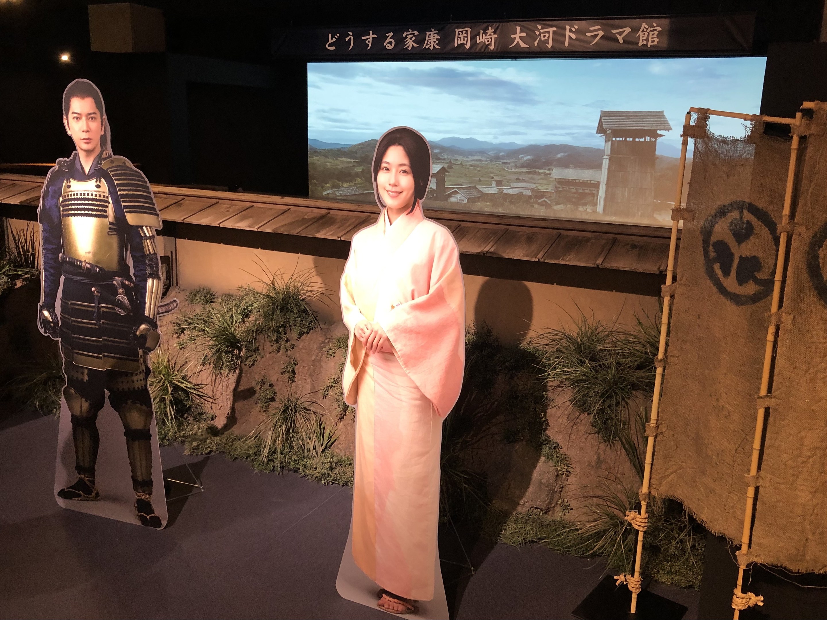 Zwei Menschen in traditioneller japanischer Kleidung - eine Frau in einem pinken Kimono und ein Mann in Samuraikleidung - lächeln gemeinsam auf einem Set mit Pflanzen, einem Bildschirm und Beleuchtungsausrüstung.