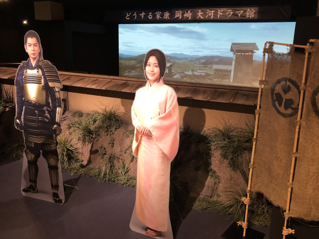 Zwei Menschen in traditioneller japanischer Kleidung - eine Frau in einem pinken Kimono und ein Mann in Samuraikleidung - lächeln gemeinsam auf einem Set mit Pflanzen, einem Bildschirm und Beleuchtungsausrüstung.