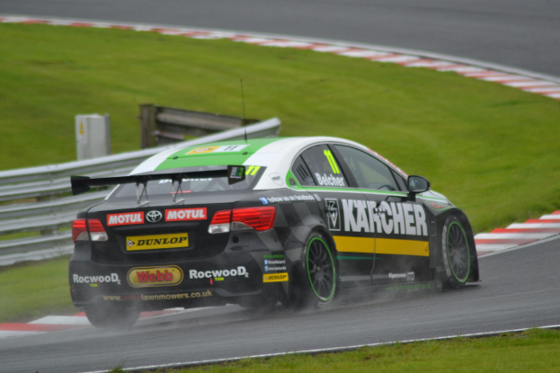 Ein Auto mit Text auf der Seite, das auf einer regennassen Rennstrecke fährt, umgeben von Gras, mit einem Geländer und einer hölzernen Konstruktion im Hintergrund.