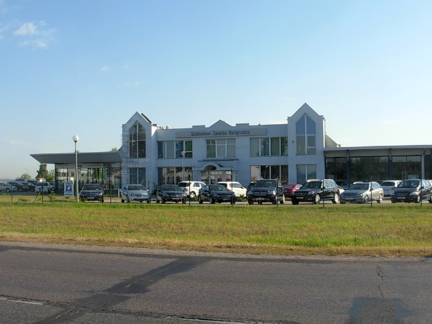 Ein Autohaus mit parkenden Autos davor, umgeben von Gebäuden, Straßeninfrastruktur, fahrenden Fahrzeugen, Bäumen und einem bewölkten Himmel.