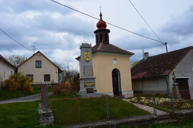 Kleine Kirche mit einem Glockenturm als Kirche des Heiligen Grabes, zentral in einem kleinen Dorf mit Häusern, einem Zaun, Gras, blühenden Pflanzen, einem Weg, Bäumen, Strommasten mit Drähten und einem bewölkten Himmel.