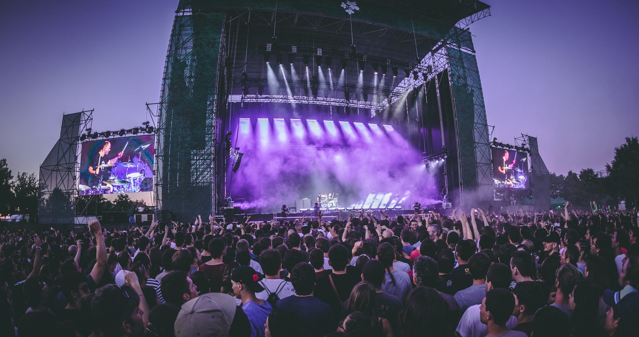 Eine große Menschenmenge auf einem Musikfestival schaut Künstlern auf einer hell erleuchteten Bühne mit Instrumenten und Bildschirmen zu, die vor einem Gebäude, Bäumen und einem klaren blauen Himmel aufgebaut sind.