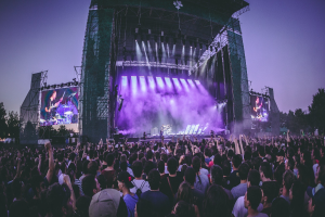 Eine große Menschenmenge auf einem Musikfestival schaut Künstlern auf einer hell erleuchteten Bühne mit Instrumenten und Bildschirmen zu, die vor einem Gebäude, Bäumen und einem klaren blauen Himmel aufgebaut sind.