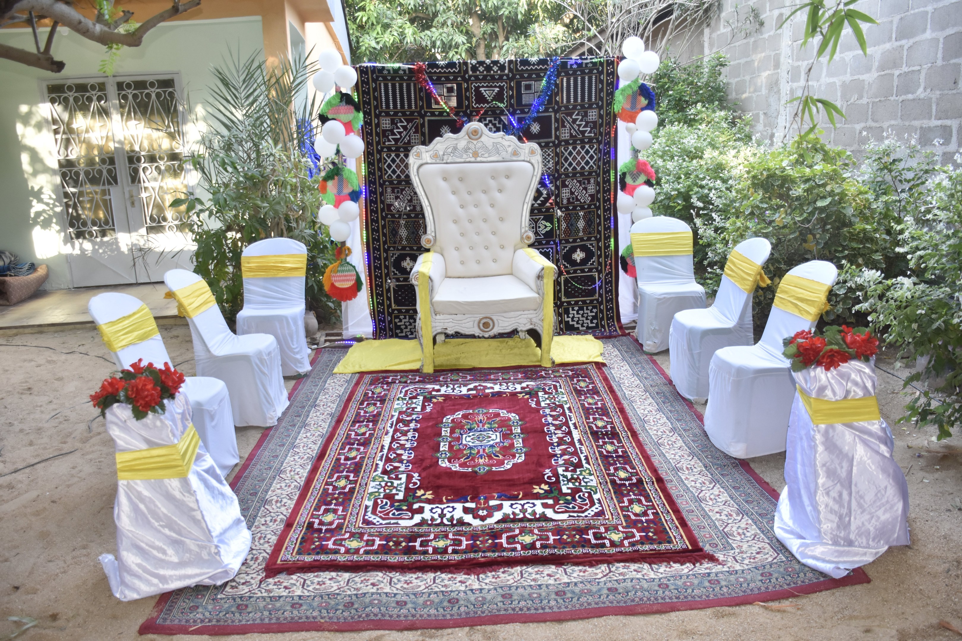 Hochzeitszeremonie-Aufbau in einem Garten mit weißen Stühlen, einem roten Teppich, Blumenbouquets, Dekorationsgegenständen, Ballons, Pflanzen, Bäumen, einem Gebäude mit Fenstern und einer Tür und einer Wand auf der rechten Seite.