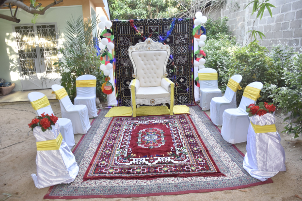 Hochzeitszeremonie-Aufbau in einem Garten mit weißen Stühlen, einem roten Teppich, Blumenbouquets, Dekorationsgegenständen, Ballons, Pflanzen, Bäumen, einem Gebäude mit Fenstern und einer Tür und einer Wand auf der rechten Seite.