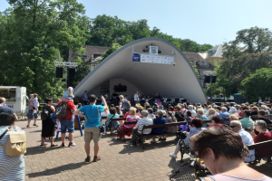 Menge auf Bänken vor einer Bühne mit Stehenden, einem Fotografen, einem Stativ, einer Fahne, Metallrahmen, Bäumen, Gebäuden und einem bewölkten Himmel bei einem Sommerpark-Konzert.