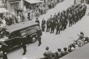 Schwarzes und weißes Bild einer Trauerprozession mit einem Leichenwagen, der den Körper eines Mannes trägt, umgeben von Menschen, einige mit Waffen, Fahrzeugen und Gebäuden mit Fenstern und Namensschildern im Hintergrund.