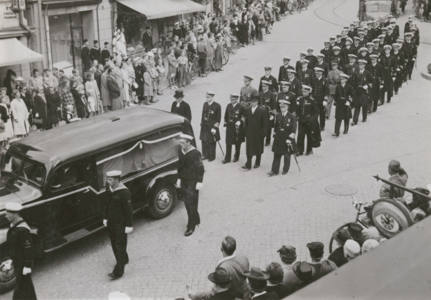 Schwarzes und weißes Bild einer Trauerprozession mit einem Leichenwagen, der den Körper eines Mannes trägt, umgeben von Menschen, einige mit Waffen, Fahrzeugen und Gebäuden mit Fenstern und Namensschildern im Hintergrund.