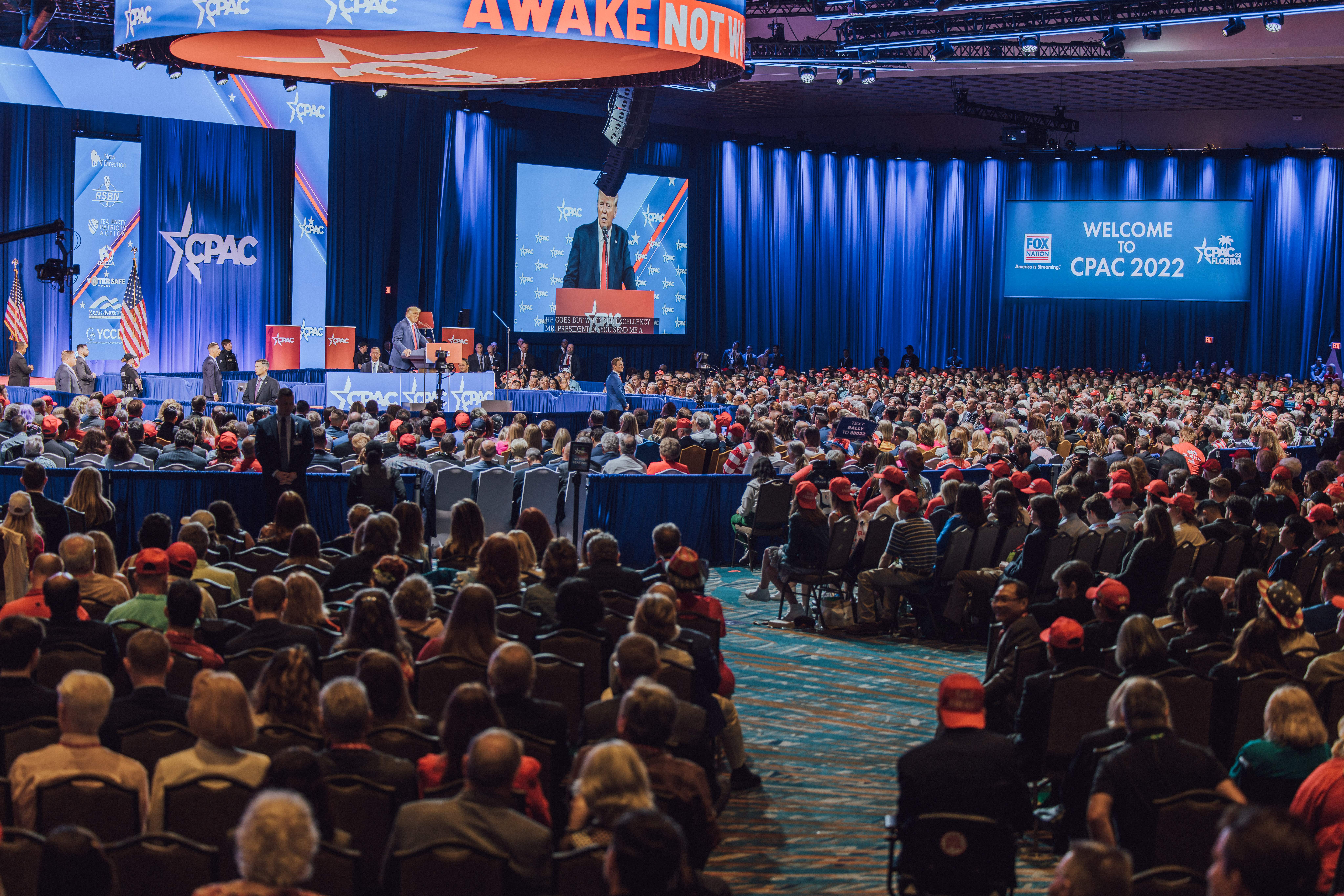 Eine große Menschenmenge sitzt auf Stühlen, die einer Bühne zugewandt sind, mit ein paar Menschen auf dem Podium und auf der linken Seite, blaue Vorhänge und ein Bildschirm im Hintergrund und eine orangefarbene Tafel oben, die das CPAC 2020-Ereignis am 25. Oktober anzeigt.