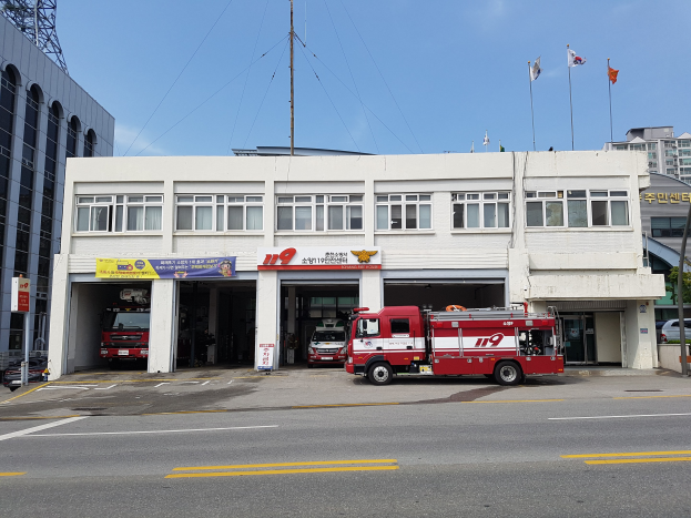 Feuerwache mit mehreren Feuerwehrautos davor geparkt, ein Gebäude mit Fenstern, Bannern und Fahnen, unter einem bewölkten Himmel mit einem Turm links.