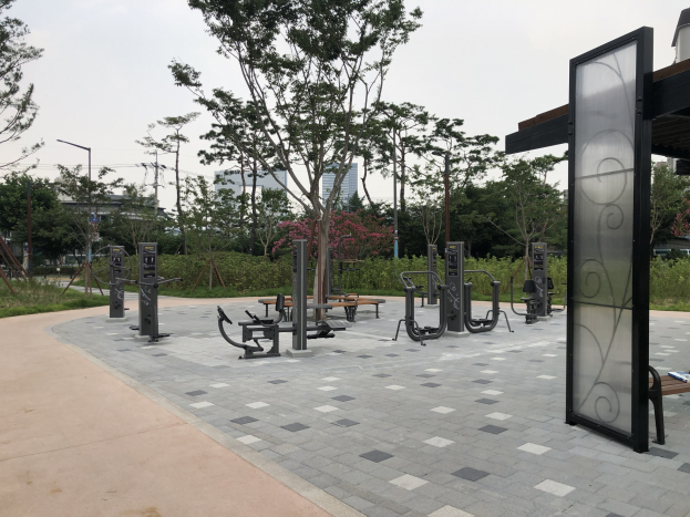 Outdoor park featuring various fitness equipment, benches, trees, plants, grass, poles, lights, wires, and buildings under a sky backdrop.