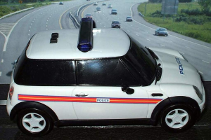 Weißes Polizeiauto mit "Polizei"-Aufschrift auf einer Straße außerhalb der Stadt, umgeben von fahrendem Verkehr, Gras und Bäumen auf beiden Seiten.