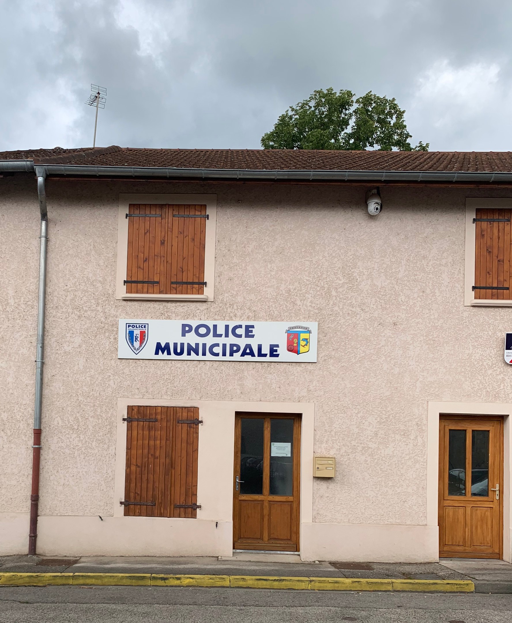 Polizeigebäude mit einem Schild, Fenstern, Türen, einer Namensplakette, einem Rohr, einem Baum und einem bewölkten Himmel im Hintergrund.
