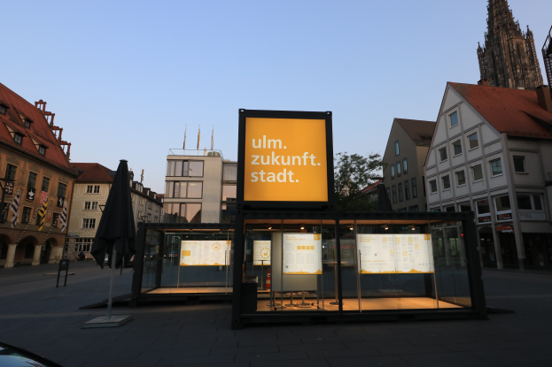 Ein Bushaltestelle in einem Stadtplatz mit einem geparkten Auto davor, umgeben von Gebäuden, Bäumen und Fahnenmasten, mit einer Texttafel im Vordergrund und einem klaren blauen Himmel im Hintergrund.