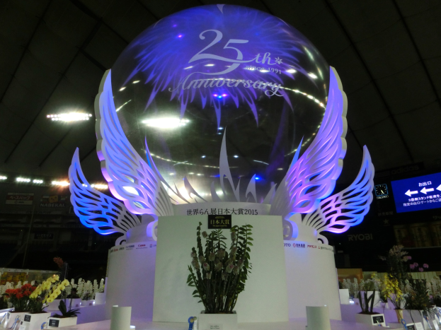 Ein großer Kristallball mit Flügeln oben drauf, umgeben von Blumenbouquets, Namensschildern und anderen Gegenständen auf dem Boden, vor einem festlichen Hintergrund mit Schildern, Lichtern und einem Bildschirm.