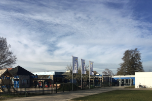 Großes Gebäude mit einem grasbewachsenen Bereich davor, umgeben von einem Metallzaun, mit einem Weg, Bäumen, Pfählen, Bannern, einem geparkten Fahrrad, nahegelegenen Häusern und einem bewölkten Himmel, identifiziert als der neue Spielplatz der Schule.