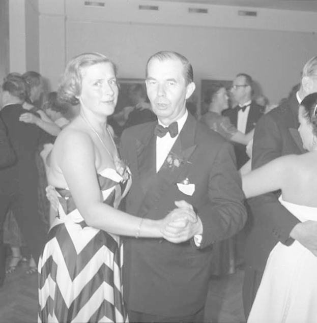 Ein Mann in einem schwarzen Blazer, weißem Hemd und schwarzen Hosen tanzt mit einer Frau in unbekannter Kleidung auf einem formellen Event, indem sie sich an den Händen halten und zur Musik schwenken, nahe einer Wand.