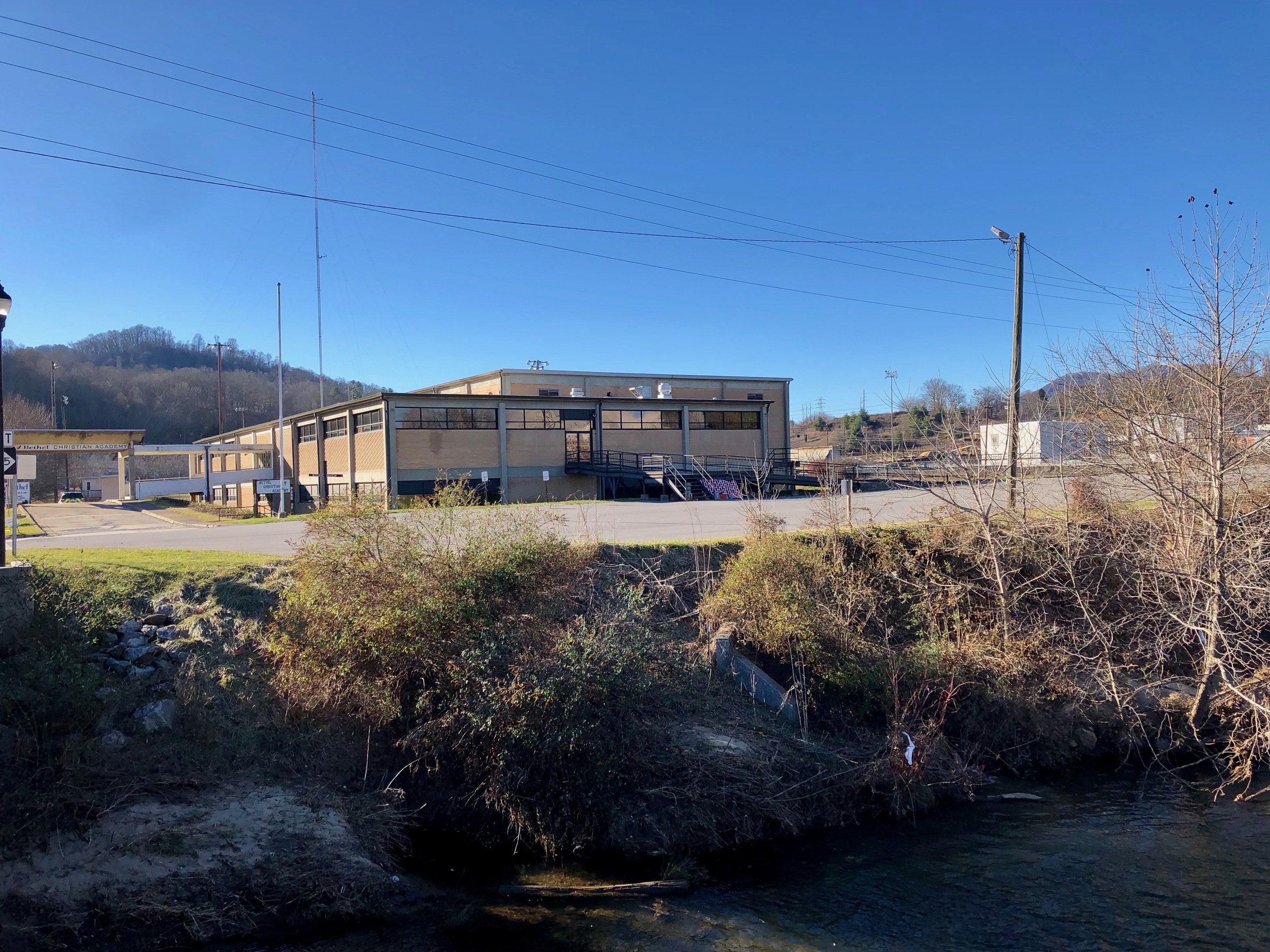 Ein Fluss durchquert eine grüne Wiese neben einem Gebäude in der Nähe von Bäumen, Pflanzen, Gras, Steinen, Straßenlaternen, Strommasten, Stromkabeln, Hügeln und einem klaren blauen Himmel, wobei das Gebäude als Standort einer geplanten Wasseraufbereitungsanlage vorgesehen ist.